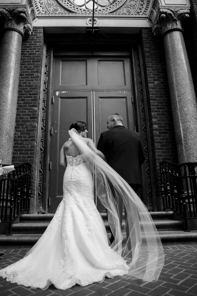 Bride and father await to enter church. Bride's veil is swept away in the wind.