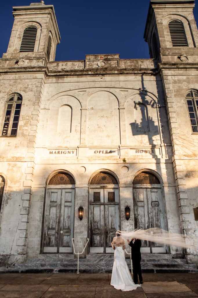 bride standing in front of Marigny Opera House with her father, her veil is swept up in the wind