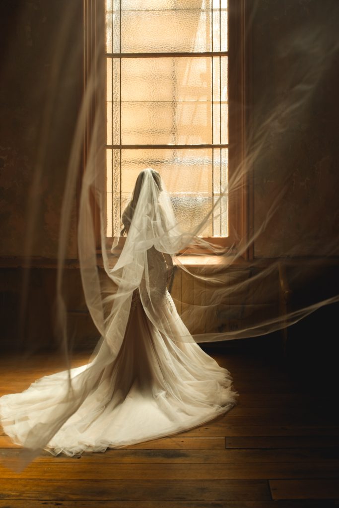 Bride's veil floats in the air creating a dramatic scene while bride looks out window at Hotel Peter & Paul Church