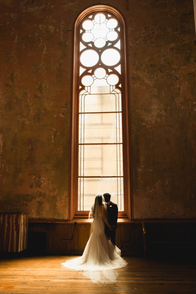 Bride and groom embrace in the light of a stained glass window